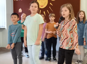 Dziewczynki i chłopcy z klasy 2b śpiewają piosenki. W tle kolorowa dekoracja z roześmianym słońcem i napisem "Bądź powodem czyjegoś uśmiechu".