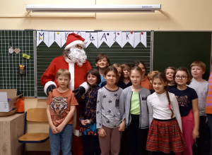 Dzieci i Mikołaj stoją w sali na tle szkolnej tablicy. Pozują do pamiątkowego zdjęcia.