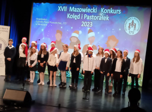 Na scenie stoją chórzyści ubrani w stoje galowe, na głowach mają świąteczne czapki. Za nimi napis XVII Mazowiecki Konkurs Kolęd i Pastorałek.