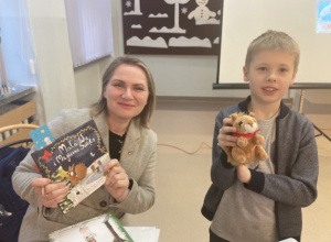 Pisarka trzyma w rękach swoją książkę pt. "Mała WU i magiczne Święta". Obok stoi uczeń klasy 2a. Trzyma w ręku wiewiórkę, bohaterkę książek Pani Pazusinskiej. Obydwie osoby uśmiechają się.