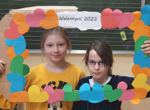 Dwie dziewczynki trzymają ramkę z napisem Walentynki 2023 umieszczonym na środku górnej listewki. Wokół ramki przyczepiono kolorowe serca.