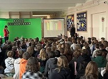 Uczniowie i nauczyciele zgromadzili się na apelu. Pani dyrektor prezentuje wyniki klasyfikacji na ekranie. Obok stoi zielona tablica z napisem osiągnięcia szkolne.