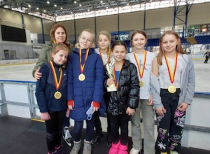 Dziewczęta z trenerką stoją przed taflą lodowiska. Zawodniczki są udekorowane złotymi medalami.