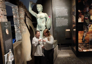 Dwie dziewczyny stoją przed rzeźbą syrenki, która trzyma w jednej ręce tarczę, w drugiej miecz.