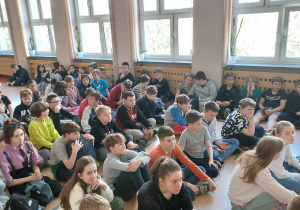 Ucziowie siedzą na podłodze szkolnego korytarza, słuchają podsumowania obchodów "Dnia Ziemi".