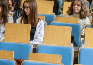 Grupa uczniów wraz z nauczycielką siedzi na auli, słuchają wykladu profesora.