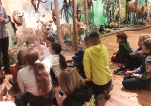 Dzieci sierdzą na podłodze. Słuchają informacji przewodnika w Muzeum Łowiectwa.