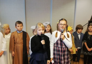 Uczniowie stoją w półkolu. Dwie dziewczynki stoją w środku i śpiewają kolędę.