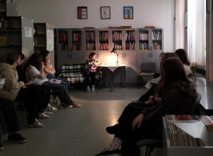 W szkolnej bibliotece uczeń klasy 8 czyta wiersz przy świetle lampki stojącej na stoliku obok niej. Przed nią siedzą uczniowie.