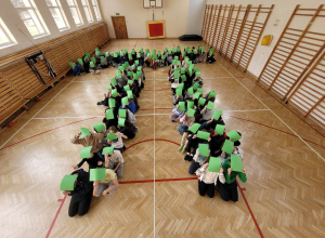Uczniowie przebywają w sali gimnastycznej. Są ustawieni w kształt liczby Pi. Nad głowami trzymają zielone kartki.