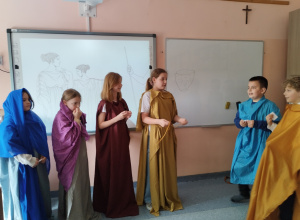 Uczniowie stoją w sali przed tablicą i odgrywają scenkę z życia starożytnych Greków. Ich stroje imitują dawny ubiór grecki.