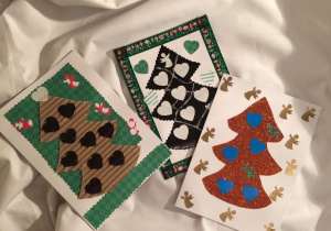 Na białym materiale rozłożone są trzy kartki wykonane przez dzieci z Domu Dziecka: pierwsza kartka przedstawia brązową choinkę z czarnymi serduszkami na zielonym tle, druga czarną choinkę z białymi serduszkami na białym tle a trzecia złotą choinkę z błękitnymi serduszkami.