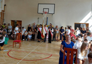 Uczniowie są zgromadzeni w sali gimnastycznej. Siedzą na ławkach. W głębi stoi poczet sztandarowy. fot. K. Sobczyk