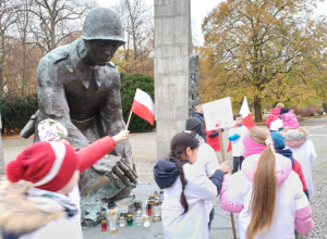 Pani w czerwonej kurtce opowiada dzieciom trzymającym flagi w białych i czerwonych koszulkach o historii pomnika Sapera.