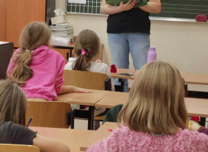 Uczniowie siedzą przy stolikach, przed nimi stoi kobieta ubrana w czarną koszulkę w rękach trzyma książkę.