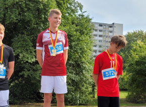 Chłopiec stoi na najwyższym podium. Ubrany jest w czerwoną koszulkę i białe, krótkie spodenki.