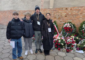 Dwóch ucznów, nauczyciel i dziewczyna stoją na tle ceglanego muru. Obok nich z prawej strony leżą wieńce. fot. Łukasz Pawłowski