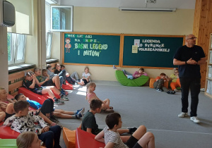 Dzieci (około 20 osób) siedzą lub leżą na kolorowych pufach (zielone, czerwone, pomarańczowe) rozrzuconych po wykładzinie. Zwrócone w stronę dorosłego mężczyzny. Nauczyciel stojący po prawej stronie zdjęcia, ubrany na czarno, opowiada dzieciom o Wiśle.