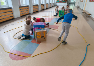 Dzieci są zaangażowane w zabawę kreatywną, wykorzystując do tego pudełka kartonowe ozdobione rysunkami i kolorowym papierem – niektóre z nich przypominają budynki z oknami. Na podłodze ułożone są kolorowe sznury, które wyznaczają granice, tworząc kształt miasteczka.