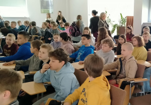 Uczniowie siedzą na krzesłach z pulpitami. Dzieci są w wieku szkolnym, ubrane w codzienne ubrania – bluzy, swetry, dresy. Niektóre z nich wydają się być skupione, inne rozmawiają między sobą lub rozglądają się po pomieszczeniu. W tle widać kilka osób dorosłych.