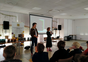 Na pierwszym planie widzimy grupę uczniów siedzących na krzesłach, skierowanych w stronę dwóch osób stojących przed ekranem projekcyjnym. Jedna z osób, kobieta w ciemnozielonej sukience i różowych butach, trzyma mikrofon i przemawia. Obok niej stoi młody mężczyzna w brązowej kurtce z naszywkami.