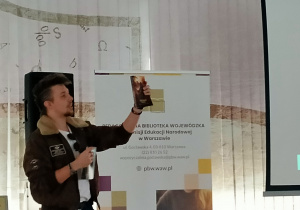 Mężczyzna ubrany jest w kurtkę typu pilotka z kołnierzem z futerkiem oraz jasny T-shirt. Trzyma w ręku książkę uniesioną w górę.