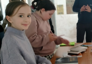 Dzieci stoją przy stole w jasnym pomieszczeniu. Na pierwszym planie znajduje się dziewczynka w jasnofioletowym swetrze, uśmiechająca się do aparatu. Obok niej druga dziewczynka w różowej bluzie z kapturem jest zajęta oglądaniem lub przeglądaniem czegoś na stole. Na blacie leżą książki, w tym jedna zatytułowana „Feel Alive”. W tle widzimy kilka innych osób, w tym dorosłego mężczyznę w okularach.