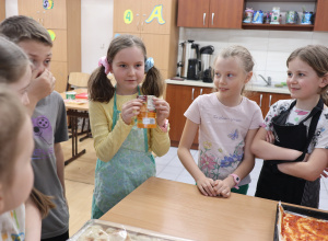 Na fotografii można zobaczyć grupę dzieci, gromadzących się wokół stołu. Jedna z dziewczynek trzyma w rękach małe opakowanie przypraw do pizzy.