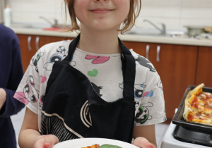 Na zdjęciu znajduje się uśmiechnięta dziewczynka w fartuchu kuchennym, trzymająca talerz z kawałkiem własnoręcznie przygotowanej pizzy. Na pizzy widoczne są liście świeżej bazylii. W tle widać kuchnię szkolną z brązowymi szafkami i zlewem. Dziewczynka wygląda na zadowoloną z efektów swojej pracy kulinarnej.