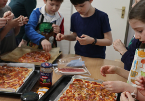 Na zdjęciu widać grupę dzieci przygotowujących pizzę w sali lekcyjnej. Dzieci stoją wokół stołu, na którym znajdują się trzy duże prostokątne blachy z ciastem do pizzy. Na pizzy widać już rozsmarowany sos pomidorowy i niektóre dodatki, takie jak szynka. Na stole leżą też różne składniki i przybory kuchenne, między innymi słoik sosu pomidorowego marki „Basilico”, opakowanie szynki dojrzewającej, biały sos w plastikowym kubeczku z łyżeczką, wałek do ciasta, opakowania foliowe i inne produkty spożywcze. Dzieci są skupione na przygotowywaniu pizzy – niektóre rozkładają składniki, inne sięgają po kolejne.