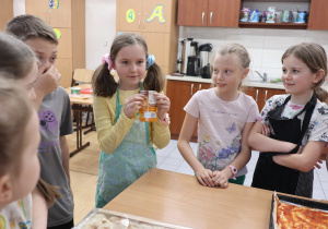 Na fotografii można zobaczyć grupę dzieci, gromadzących się wokół stołu. Jedna z dziewczynek trzyma w rękach małe opakowanie przypraw do pizzy. Dzieci przygotowują się do wspólnego wąchania i poznawania przypraw.