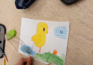 Na ilustracji znajduje się rysunek kurczaka w krajobrazie trawy, chmurki i wody. Ilustracja nie jest skończona, dziewczyna jest w trakcie jej tworzenia.
