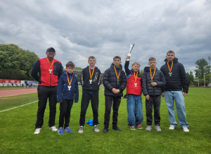 Na zdjęciu widoczna jest siedmioosobowa grupa stojąca na zielonym boisku lekkoatletycznym. Sześciu chłopców i jeden dorosły mężczyzna (trener) pozuje do zdjęcia po zawodach.