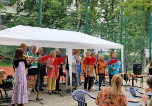 Na zdjęciu widać scenę na świeżym powietrzu zorganizowaną pod białym namiotem. Przed namiotem występuje grupa seniorów, która śpiewa lub gra na instrumentach – skrzypcach i flecie. Na scenie znajduje się statyw z mikrofonem oraz czerwone pulpity z nutami. W tle widać ogrodzenie boiska sportowego. Publiczność siedzi przy plastikowych stołach i krzesłach – na pierwszym planie widzimy kobietę w bluzce z wzorem rudych kotów.