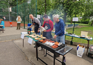Na zdjęciu widać scenę z grilla plenerowego. Trzech mężczyzn przygotowuje jedzenie na dużym grillu – pieką się na nim różne rodzaje kiełbasek. Dwóch z mężczyzn nosi fartuchy z logo programu „MasterChef”. Na stole obok grilla znajdują się papierowe talerze i butelki z przyprawami. Przyklejone kartki z cenami informują o kosztach porcji: „Kiełbasa + bułka” za 10 zł.