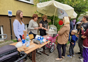 W centrum znajduje się stoisko z watą cukrową — maszyną do jej robienia obsługują dwie kobiety, z których jedna właśnie nawija watę na patyk. Nad maszyną rozłożony jest parasol z logotypem. Po lewej stronie znajduje się stół z akcesoriami i składnikami do produkcji waty: patyczkami, butelką wody, cukrem i innymi produktami. Za stołem stoją trzy kobiety. Po prawej stronie zdjęcia ustawiła się kolejka chętnych – dzieci i dorośli czekają na swoją porcję słodkiej przekąski.