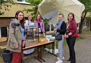 Na zdjęciu widzimy pięć kobiet stojących przy stoisku z maszyną do robienia popcornu. Jedna z kobiet jest zakonnicą w białym habicie. Stół jest ustawiony na chodniku, obok budynku z żółtą elewacją. Na stole znajduje się maszyna do popcornu, miski z gotowym popcornem, kubeczki oraz przyprawy. Nad stołem rozłożony jest duży biały parasol, który chroni stoisko.