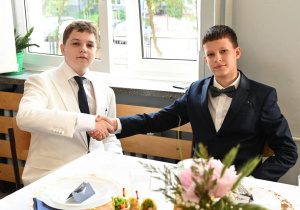 Na zdjęciu widać dwóch młodych chłopców ubranych elegancko w garnitury, którzy podają sobie ręce na powitanie. Chłopiec po lewej stronie ma na sobie biały garnitur z ciemnym krawatem, a chłopiec po prawej granatowy garnitur z muszką. Siedzą przy odświętnie nakrytym stole – na talerzach znajdują się przystawki, a na środku stołu bukiet kwiatów. W tle widać jasne okno z zieloną rośliną na parapecie, przez które wpada naturalne światło.