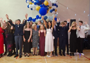 Na zdjęciu widzimy dużą grupę młodych osób w szeregu w sali gimnastycznej. Wszyscy są elegancko ubrani. Dziewczęta mają na sobie sukienki (w kolorach takich jak czerwień, czerń, bordo i biel), a chłopcy garnitury lub marynarki. Kilka osób unosi ręce w górę. W tle widać dekorację z balonów w kolorach biały, niebieski, granatowy i złoty, a po prawej stronie stoi DJ z aparaturą oraz kolorowe światła dyskotekowe,