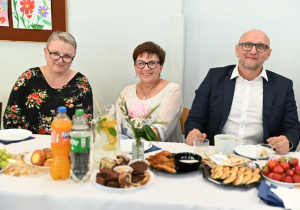 Trzech nauczycieli siedzi przy stole nakrytym. Wszystkie osoby są uśmiechnięte. Osoba po lewej, to kobieta w okularach z krótkimi, siwymi włosami, ubrana w czarną bluzkę w kolorowe kwiaty. Siedzi z lekkim uśmiechem i przechyloną głową. Osoba po prawej, to mężczyzna w okularach, ubrany elegancko w biały koszula i ciemna marynarka. W środku siedzi kobieta w okularach, krótkie brązowe włosy, biała bluzka.