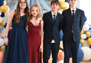 Na zdjęciu widzimy czworo elegancko ubranych młodych osób, pozujących wspólnie na tle dekoracyjnego łuku z balonów w kolorach granatowym, białym, złotym i brzoskwiniowym, z tiulową, błękitną wstęgą. Dwie dziewczyny ubrane są w długie suknie – jedna w granatową, druga w bordową – natomiast chłopcy mają na sobie klasyczne garnitury.