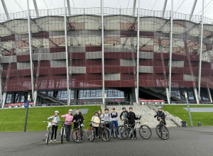 Grupa uczniów stoi z rowerami. W tle znajduje się Stadion Narodowy w Warszawie.