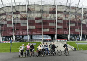 Grupa uczniów stoi z rowerami. W tle znajduje się  Stadion Narodowy w Warszawie.