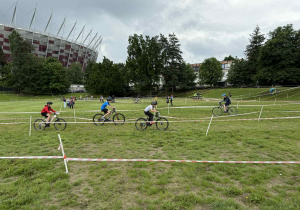 Grupa rowerzystów bierze udział w plenerowym wydarzeniu kolarskim na trawiastym terenie. Teren wyścigu jest oznaczony białą taśmą, która wyznacza trasę dla uczestników. W tle znajduje się Stadion Narodowy w Warszawie. Na trasie widać kilku rowerzystów jadących po wyznaczonej ścieżce, a także osoby stojące.