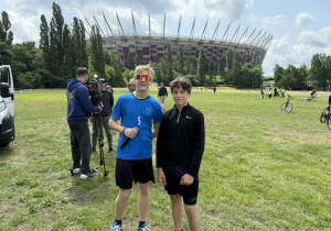 Dwie osoby stoją na trawiastym terenie, na tle stadionu. W tle znajduje się stadion o unikalnym wyglądzie, z czerwono-białym wzorem na elewacji i wysokimi masztami wystającymi z dachu. Wokół widać kilka innych osób, z których część filmuje lub robi zdjęcia. Na pierwszym planie i w tle znajdują się również rowery i drzewa.