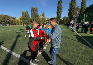 Na zdjęciu widać moment wręczania medali młodym zawodniczkom na boisku sportowym. Dziewczyny stoją w szeregu, ubrane w czerwono-czarne stroje sportowe, a mężczyzna w dżinsowej kurtce podaje jednej z nich medal z kolorową wstążką.