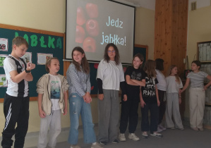 Dzieci stoją w rzędzie. Za nimi znajduje się ekran z wyświetlonym napisem „Jedz jabłka!” oraz tło z obrazkiem czerwonych jabłek. Na tablicy obok widnieje napis „Jabłka” i kilka plakatów o tym owocu.