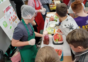 Uczniowie zgromadzili się wokół stołów, na których przygotowano różne potrawy i napoje. Kilka osób w fartuchach i czepkach kucharskich obsługuje stoisko, podając jedzenie i napoje innym. Na stole można zauważyć m.in. kawałki arbuza, dzbanki z sokiem oraz kubeczki.