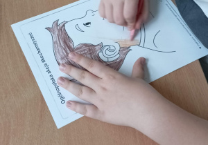 Uczeń siedzi przy stoliku i koloruje rysunek przedstawiający chłopca z aparatem słuchowym lub implantem ślimakowym.