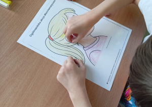 Dziewczynka w białej koszulce siedzi przy stoliku i wykleja plasteliną aparat słuchowy widoczny na kolorowance. Na stoliku leżą przybory szkolne.